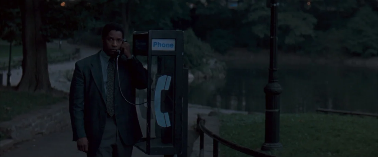 The Pelican Brief (1993) movie still: medium — A man stands next to a lone outdoor phone booth at the edge of a park. He speaks into the receiver…