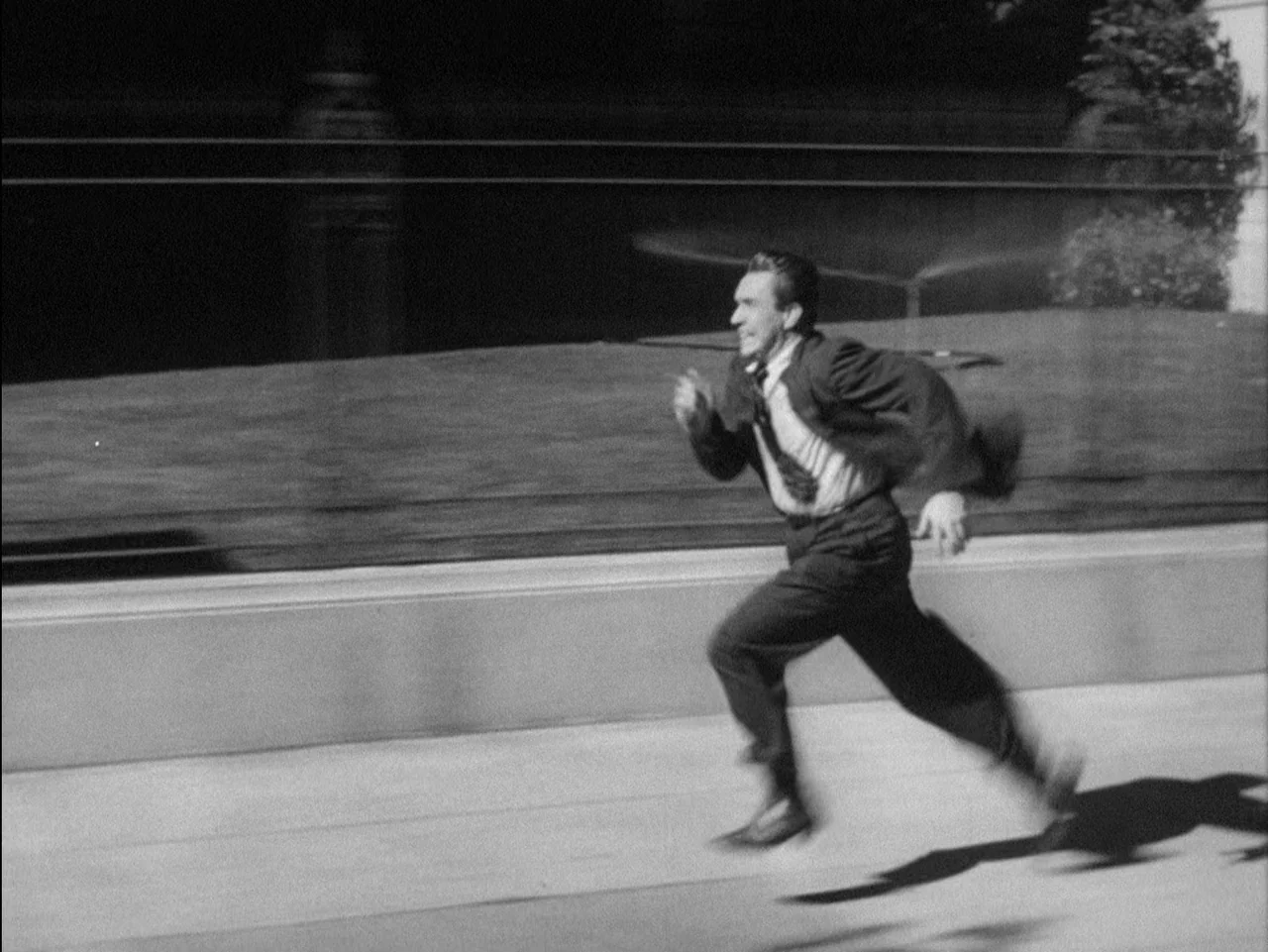 D.O.A. (1949) movie still: fullbody — A man in a suit sprints across a concrete pathway in a park. He moves laterally across the frame with motion…