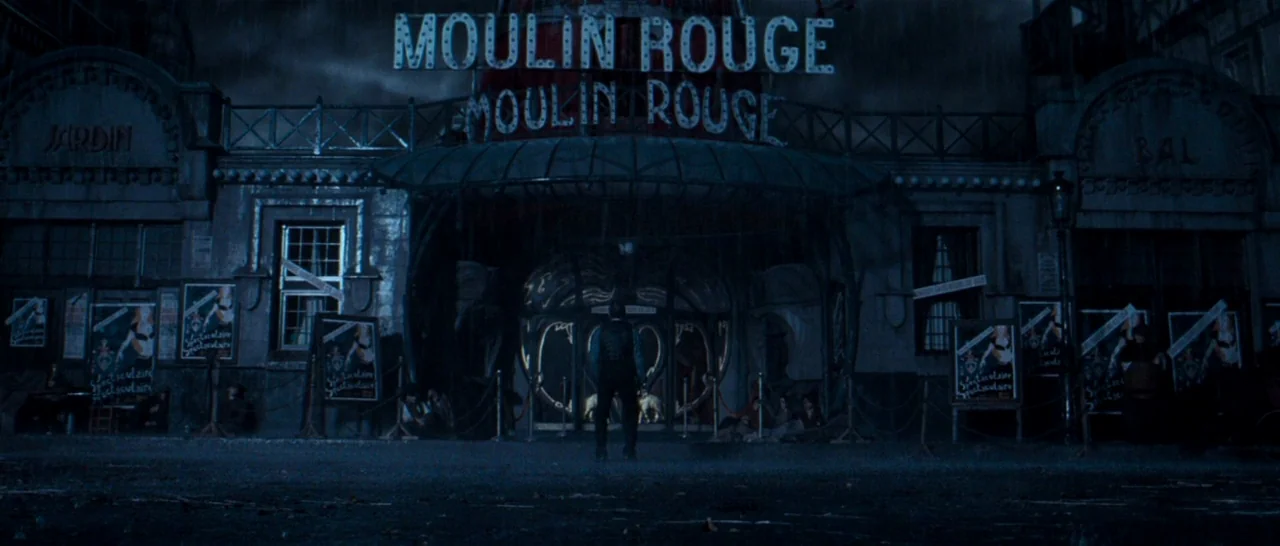 Moulin Rouge! (2001) movie still: wide — The exterior of the Moulin Rouge at night features dark architecture and illuminated signage. A lone man…