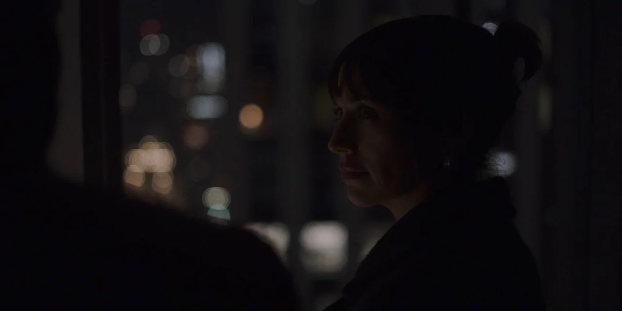 The Sound of Silence movie still: closeup — An interior space at night overlooks a blurry city skyline through a glass window. A woman stands in…