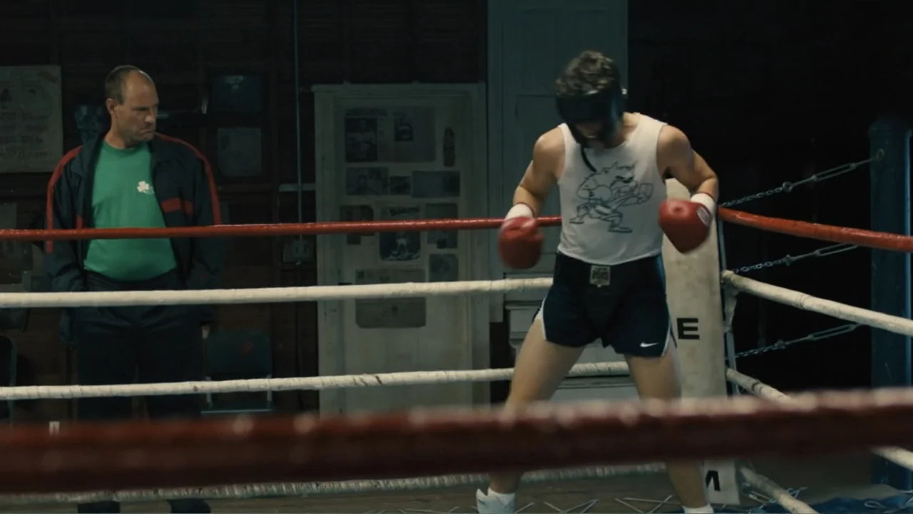 Bleed for This (2016) movie still: medium — An aging interior boxing gym with a ring featuring red and white padded ropes. A young boxer in training…