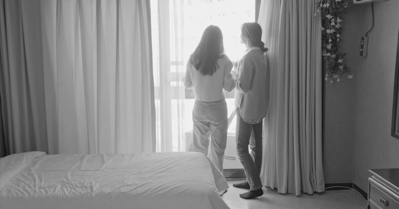 Hotel By The River movie still: fullbody — A spare hotel room features heavy curtains flanking a large floor-to-ceiling glass pane. Two women stand…