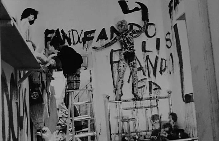 Fando y Lis (1968) movie still: medium — A stark white room is covered in large black painted letters and scattered dolls. A person paints on the…