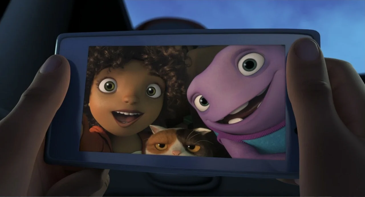 Home (2015) movie still: closeup — A handheld tablet screen displays a selfie of a girl, a purple alien, and a cat. The device is being held inside…