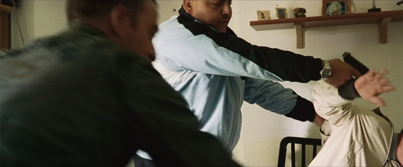 GOMORRAH (2023) movie still: medium — A residential room features a plain white wall with a small shelf holding framed photos. A man in a blue track…