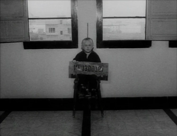 Even Dwarfs Started Small (1970) movie still: medium — An interior space with plain light-colored walls and two windows overlooking a street. A man…