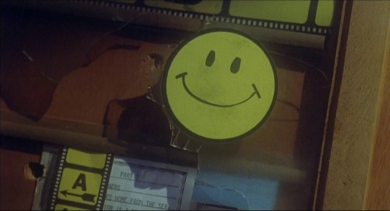The Howling (1981) movie still: closeup — A dusty glass display cabinet contains a yellow smiley face sticker and film strips. The glass surface…
