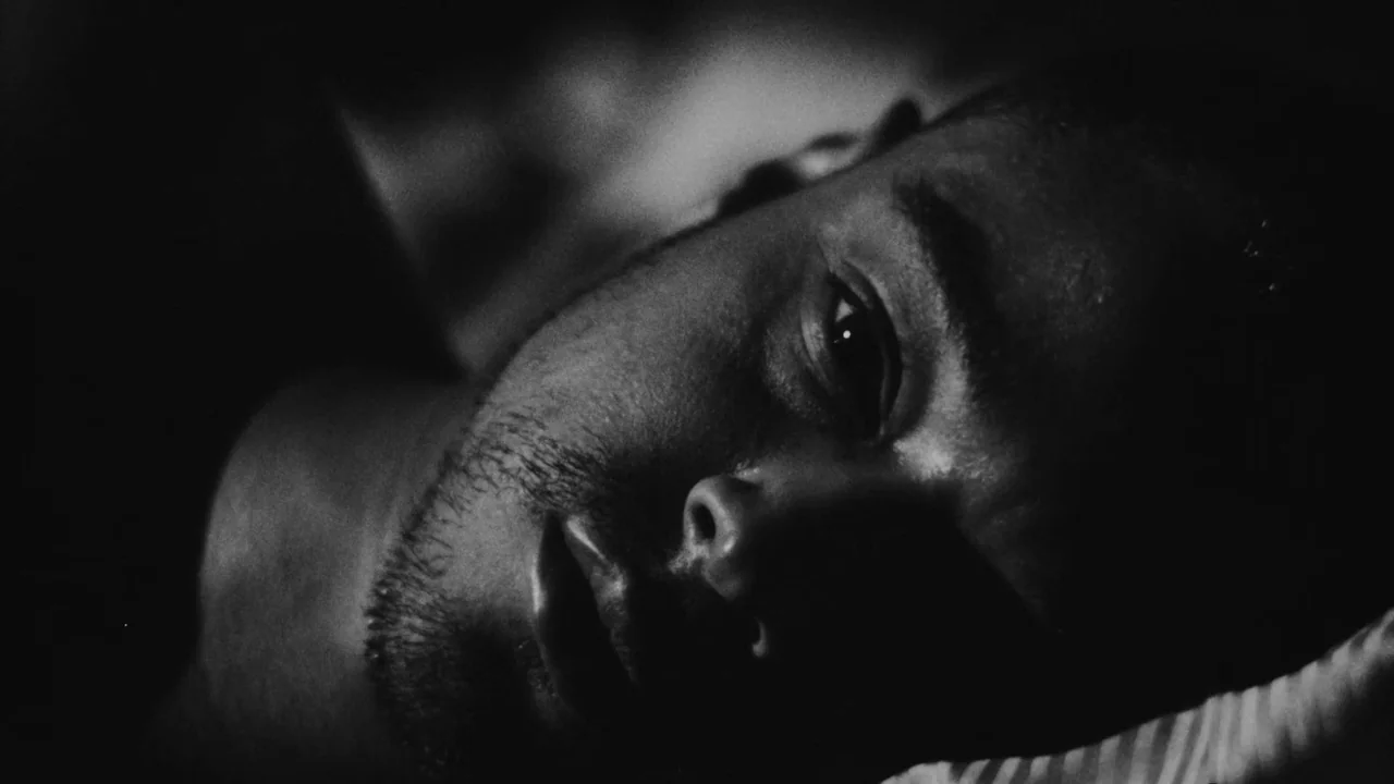 Divinity (2023) movie still: closeup — A man's face lies in shadow on a textile surface in a dark room. The man rests on a pillow while looking…