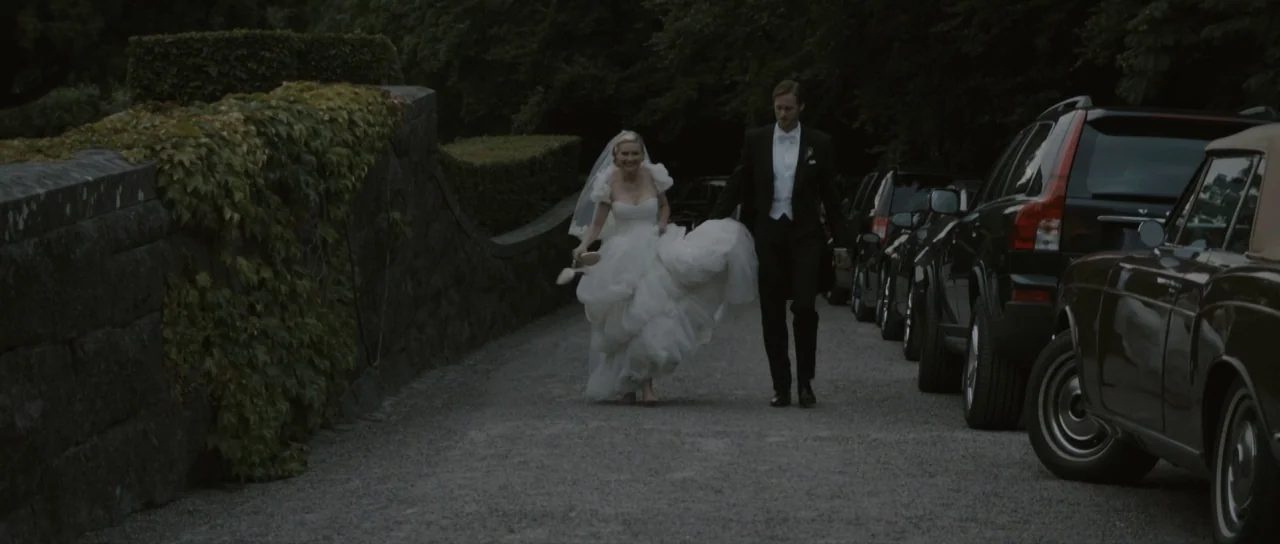 Melancholia (2011) movie still: fullbody — A gravel driveway lined with a stone wall covered in vines and parked cars. A bride holding her shoes and…