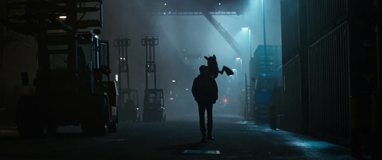 Pokémon Detective Pikachu movie still: wide — A dark, foggy shipping terminal at night is illuminated by tall, cool-toned streetlights. A man walks…