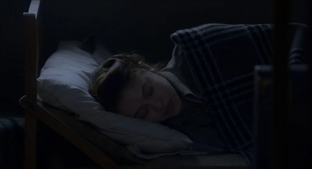 The Innocents (2016) movie still: closeup — A person rests in a dark space on a simple wooden bed frame. The subject is asleep, partially covered by…