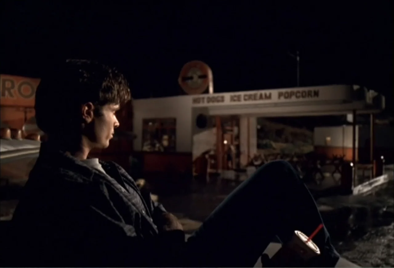Black Cat Run (1998) movie still: medium — A night exterior view features a roadside snack stand with illuminated menu signage. A young man sits in…
