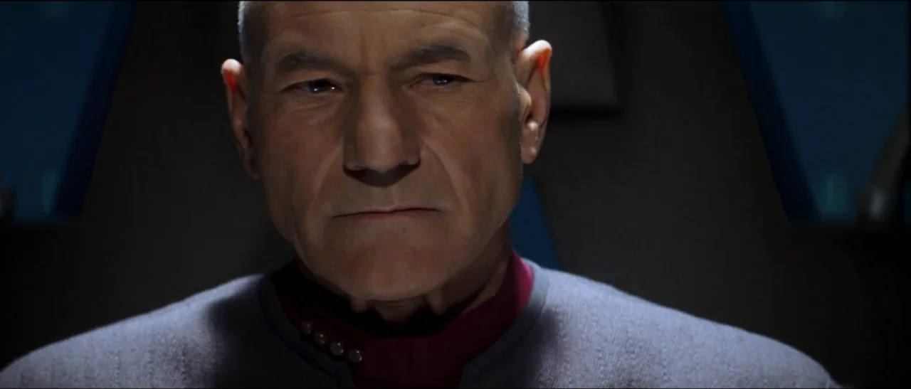 Star Trek: Nemesis (2002) movie still: closeup — A dimly lit interior station featuring dark walls and subtle technical geometric paneling. An older…