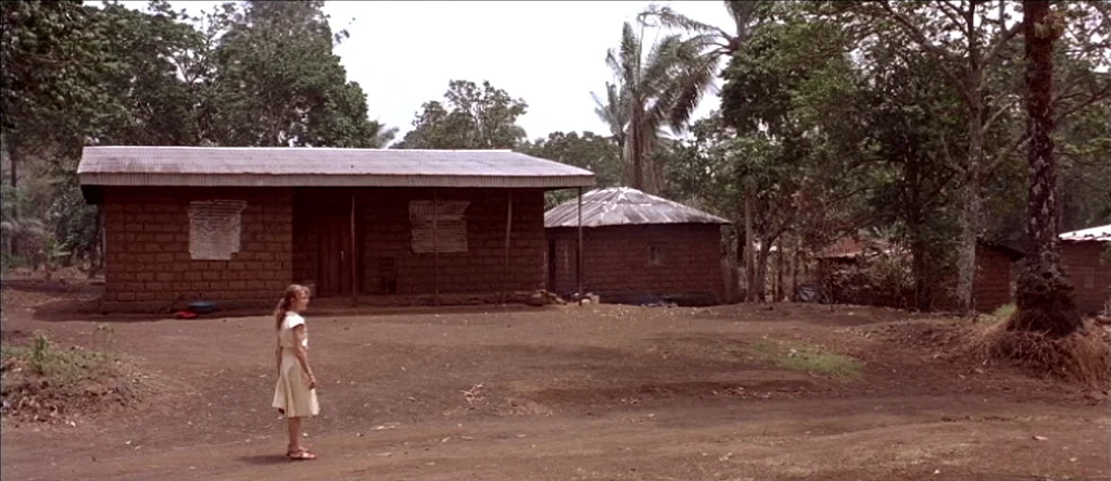 White Material (2009) movie still: wide — A rural setting with several brick dwellings under a canopy of tropical trees. A lone girl stands on the…