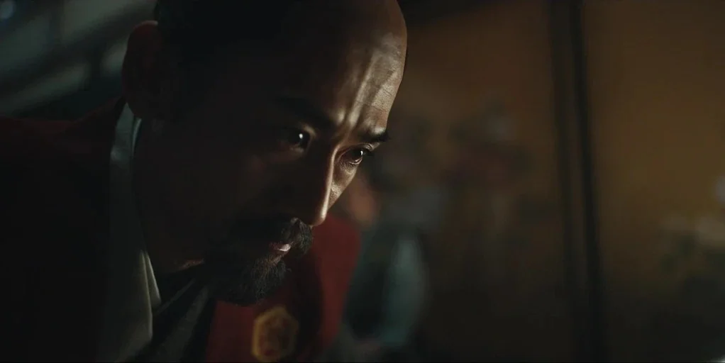 Shogun (2024) movie still: closeup — A dimly lit room with traditional Japanese architectural details visible in the soft background. An older man…