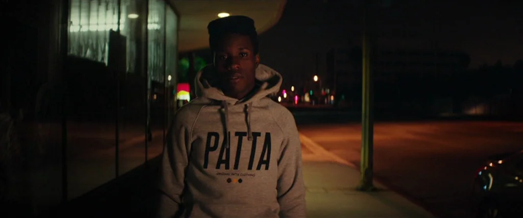 Dope (2015) movie still: medium — A dark city street at night features a building with large glass windows. A young man wearing a grey hooded…