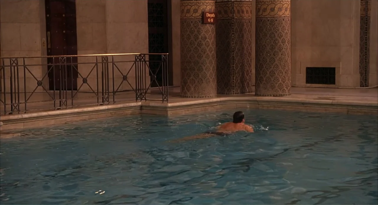 Scoop (2024) movie still: wide — An indoor swimming pool with ornate decorative columns and marble tiling walls. A lone man swims through the center…