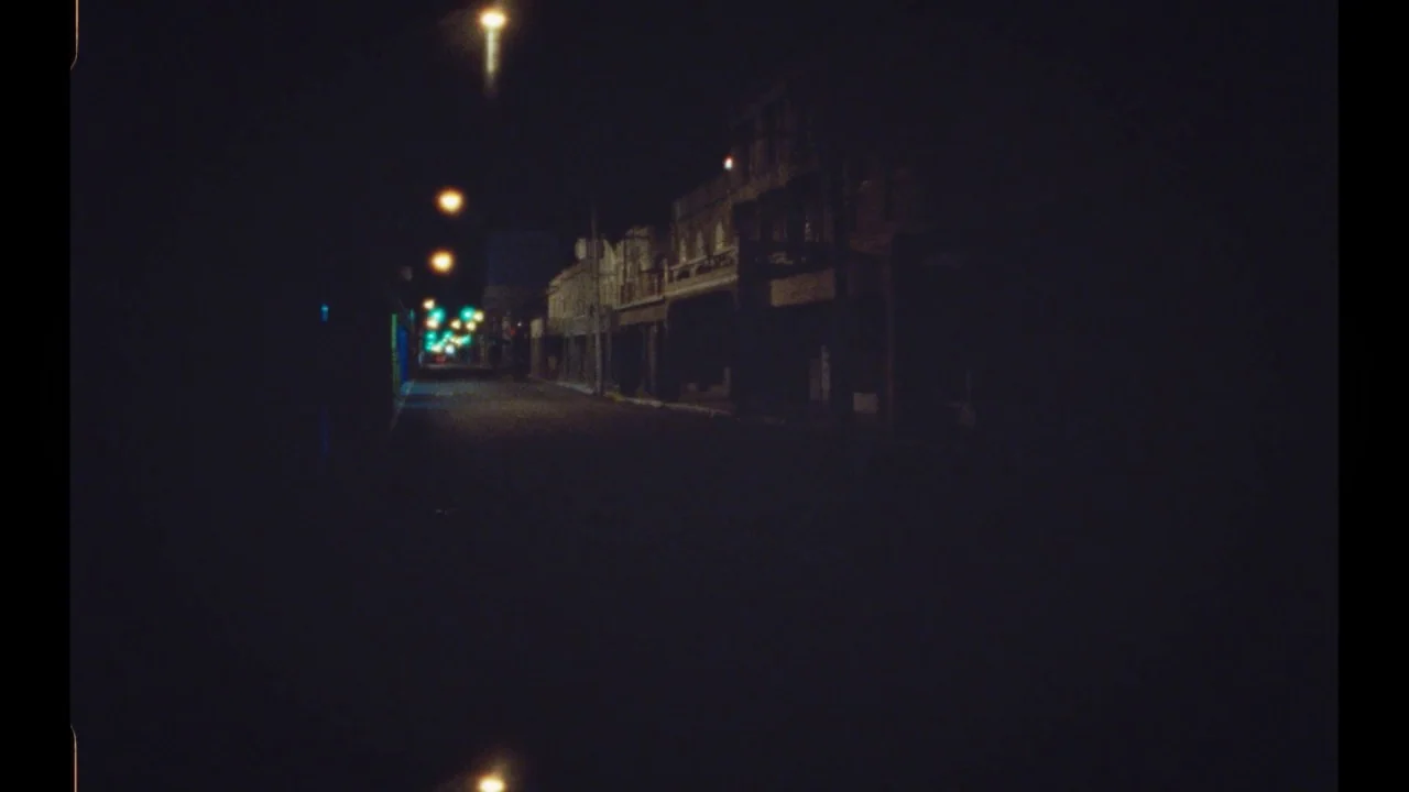Black Mother (2018) movie still: establishing — A deserted street at night features silhouetted buildings along the dark pavement. Rows of dim…