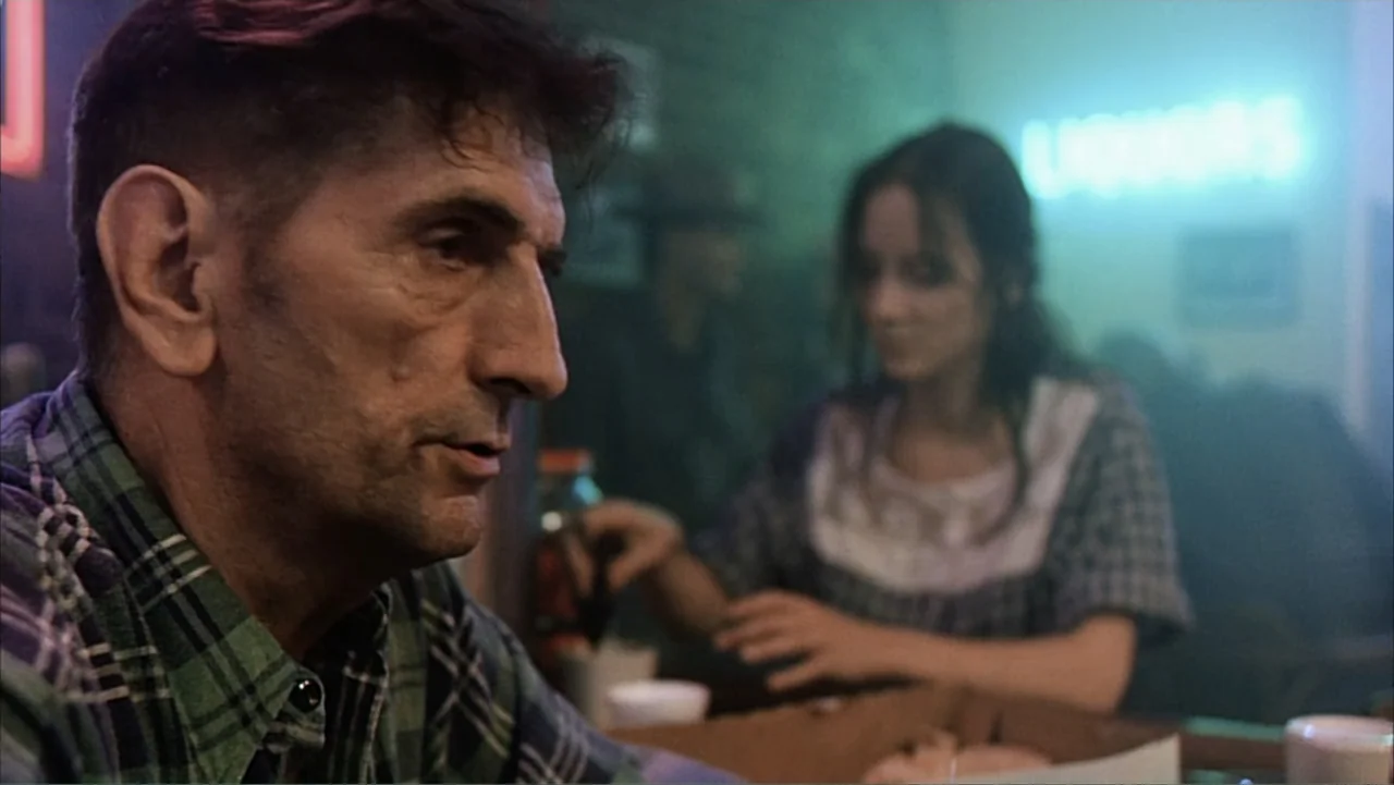 Against the Wall (1994) movie still: medium — A dimly lit diner features a glowing cyan liquor store sign in the background. An older man in a…