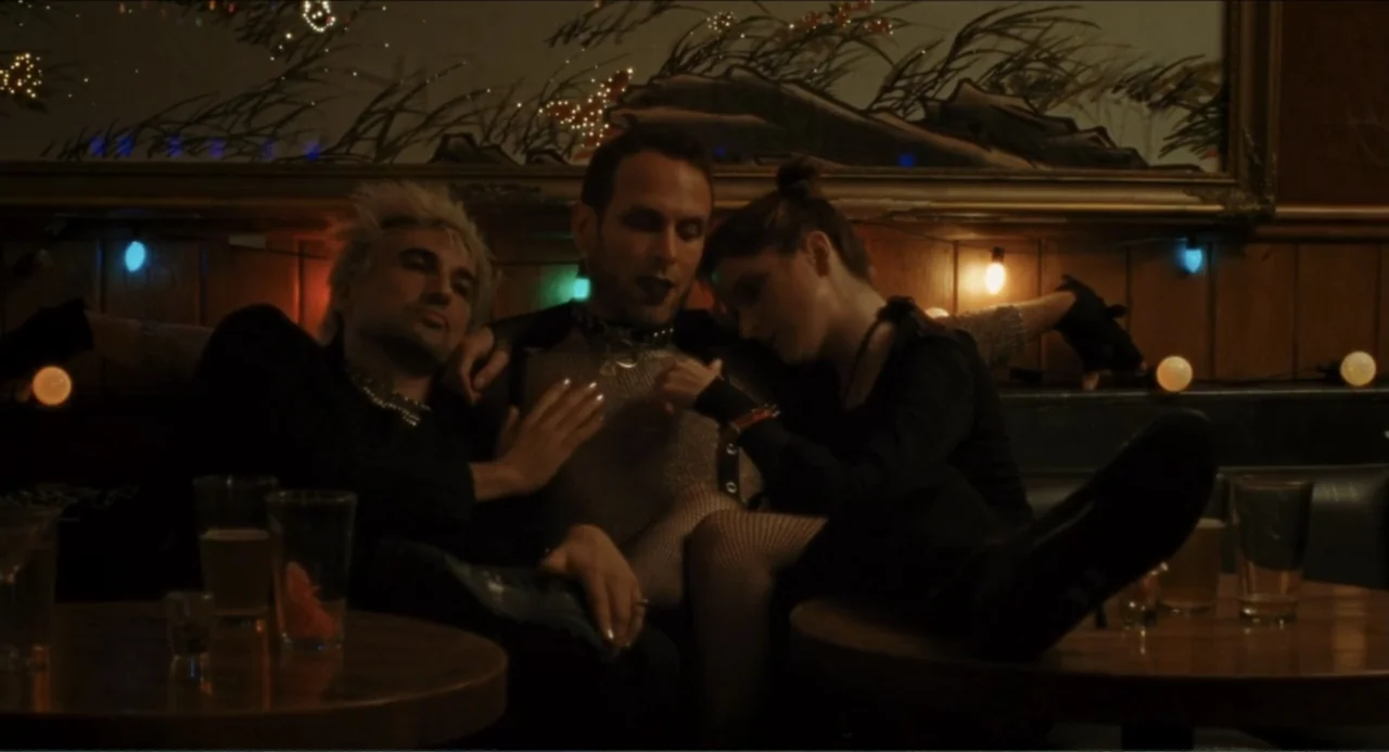 A Different Man (2024) movie still: medium — A dimly lit tavern booth features wood paneling and decorative string lights. Three individuals are…