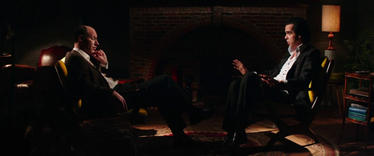 20,000 Days on Earth (2014) movie still: wide — A dimly lit lounge featuring a brick fireplace and mid-century modern furniture. Two men sit in…