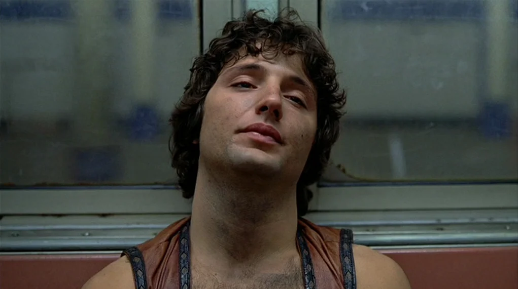 The Warriors (1979) movie still: closeup — Interior of a subway car featuring metal paneled walls and a window. A young man sits directly in front…