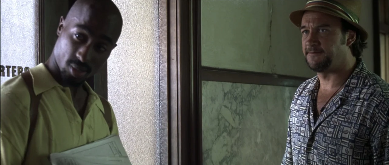 Gang Related (1997) movie still: medium — A dimly lit indoor corridor with textured walls and a wooden door frame. Two men stand together in the…