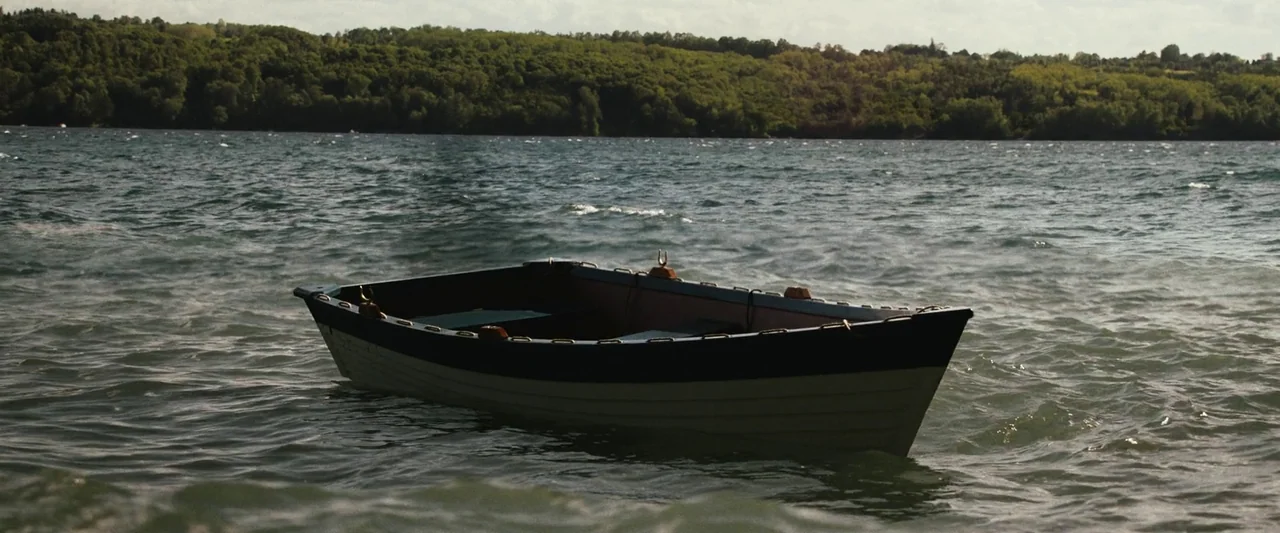 The Night House (2020) movie still: wide — A small rowboat sits empty on the surface of a rippling lake. A dense green treeline lines the distant…