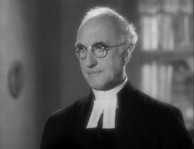 Music in Darkness (1948) movie still: closeup — An interior space features an elderly man wearing a black clerical outfit and collar. He stands…