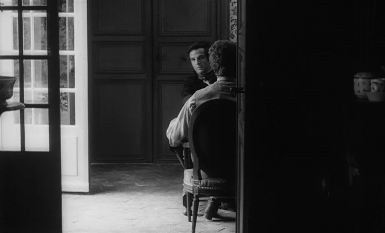 The Wild Child (1970) movie still: medium — A dark dining room with traditional wood paneling is partially lit by a nearby doorway. Two men sit at a…