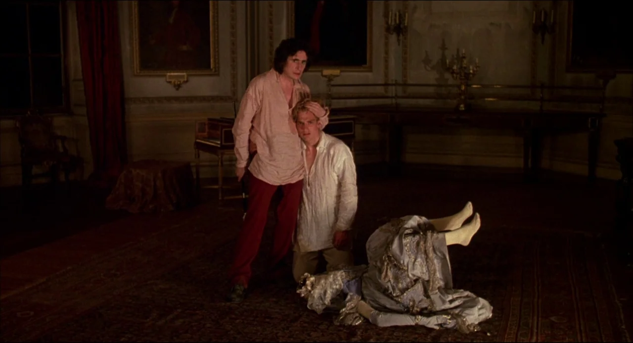 Gothic (1986) movie still: fullbody — An opulent, dimly lit room with ornate framed paintings and wood-paneled walls. Two men hover over a pile of…