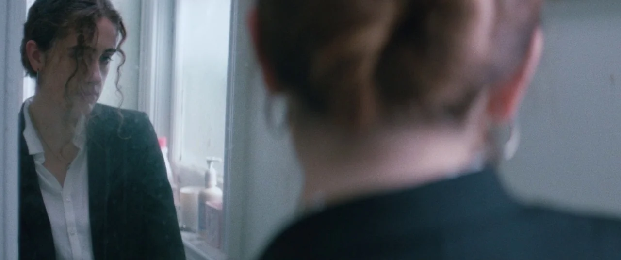 Shiva Baby (2020) movie still: medium — A woman stands in front of a mirror in a light-filled residential bathroom. She gazes at her own reflection…
