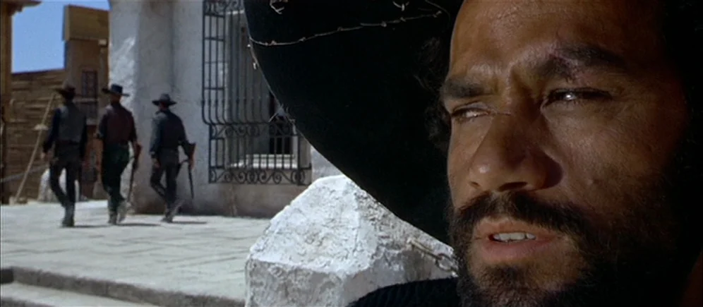 For A Few Dollars More (1965) movie still: closeup — A sunlit stone plaza sits in front of a white building with iron bars. Three men in cowboy…
