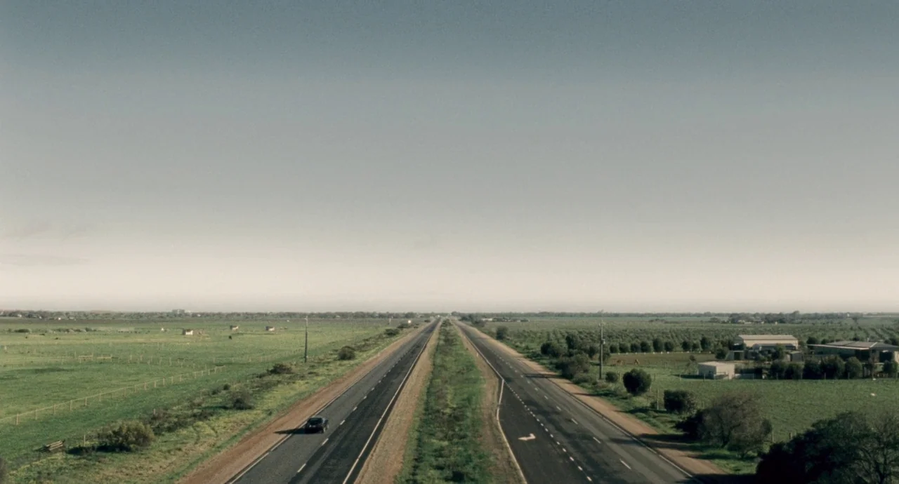 Snowtown (2011) movie still: establishing — An aerial view shows a long, straight dual-carriageway cutting through flat, expansive rural fields. A…
