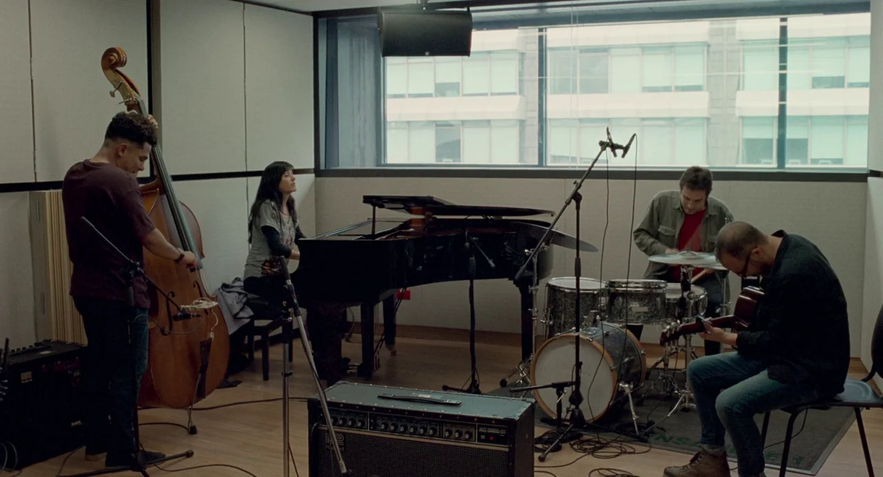 Memoria (2021) movie still: wide — A soundproof room with acoustic wall panels contains a grand piano and instruments. Four musicians play jazz…