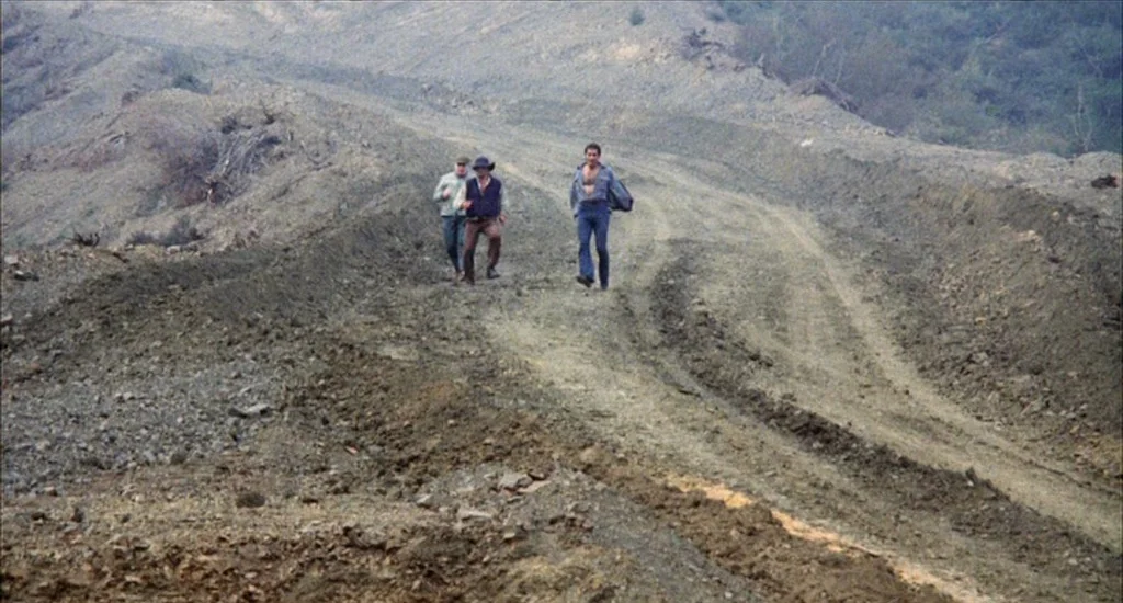 A Woman Under the Influence (1974) movie still: wide — A rugged dirt construction road winds through a barren, gray-toned hilly landscape. Three men…