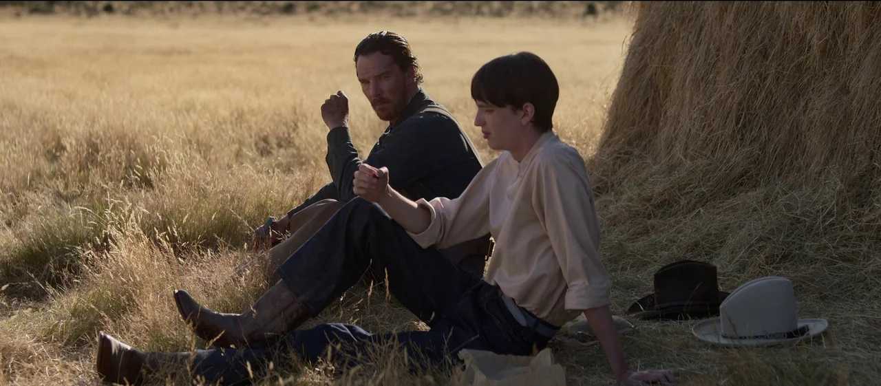 The Power of the Dog (2021) movie still: medium — A dry field with tall golden grass contains a large stack of hay. Two men sit on the ground next…