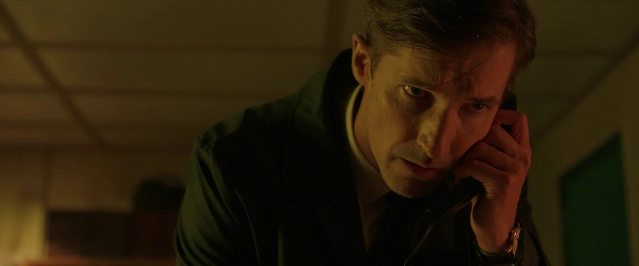 Oddity (2024) movie still: closeup — A dimly lit room with drop ceiling tiles and industrial furniture visible in shadows. A man in a dark coat…