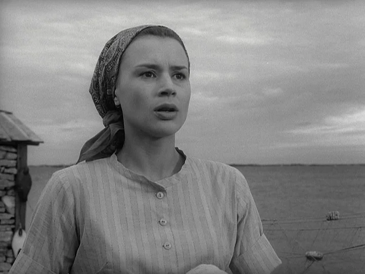 Through A Glass Darkly (1961) movie still: medium — A coastal setting with a cloudy sky and a stone structure in the background. A woman in a…