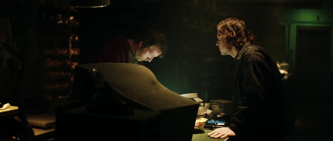 American Animals (2018) movie still: medium — A dimly lit tech workspace features a bulky monitor providing the main light source. Two men stand…