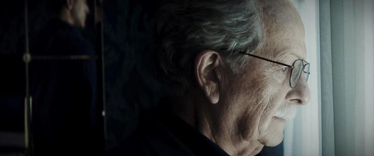 La Llorona (2019) movie still: closeup — A dimly lit room interior features a person facing toward a window with sheer curtains. The elderly man…