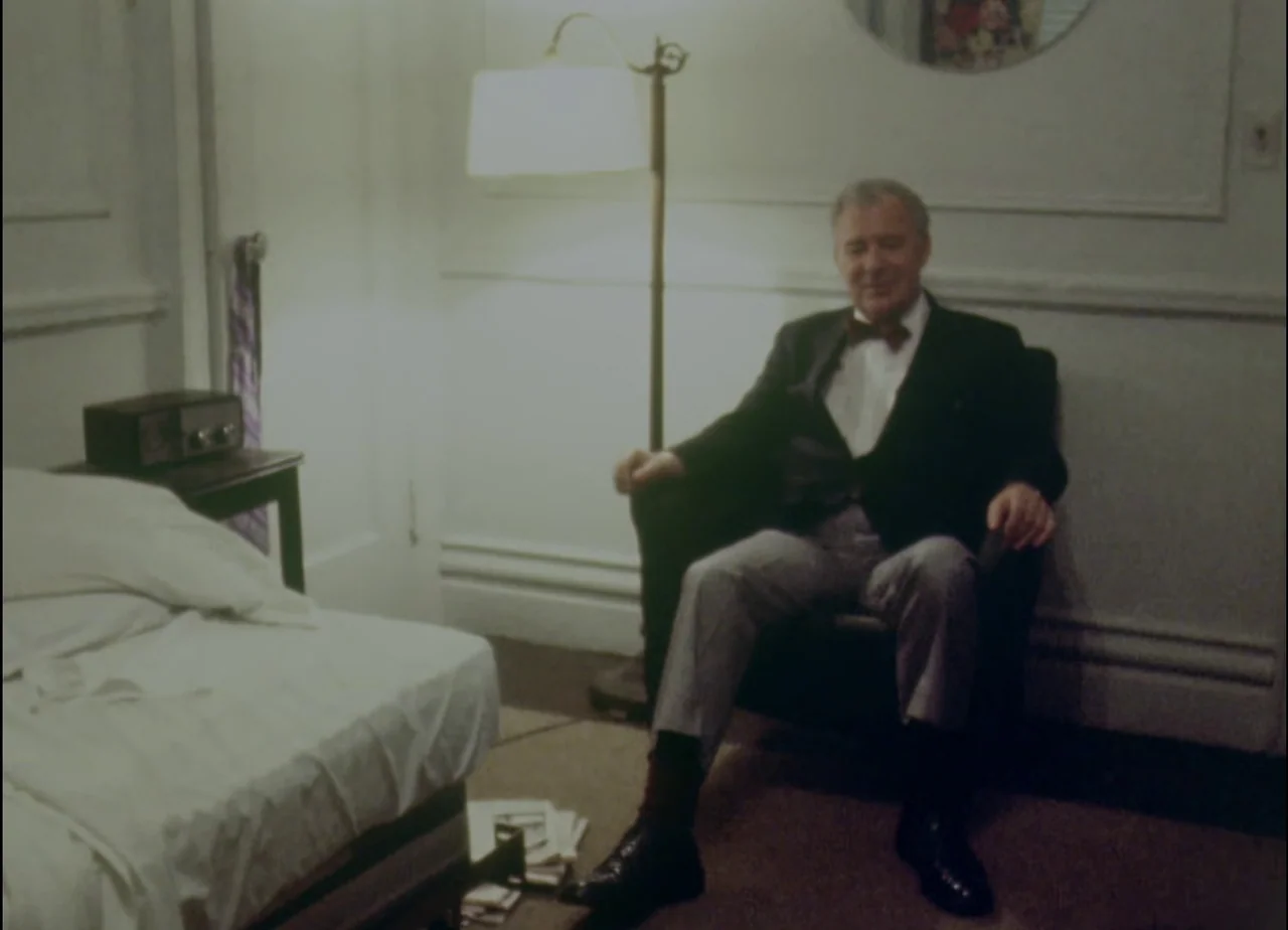 Hotel Monterey (1973) movie still: fullbody — A modest hotel room features white walls, a bed, and a floor lamp. An elderly man wearing a tuxedo…