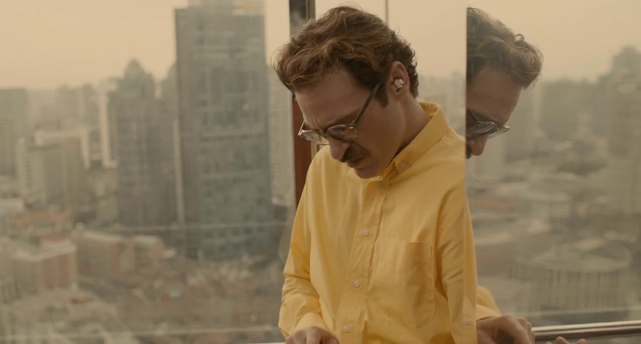 Her (2013) movie still: medium — A high-rise interior space features a large window overlooking a dense city skyline. A man wearing a yellow shirt…