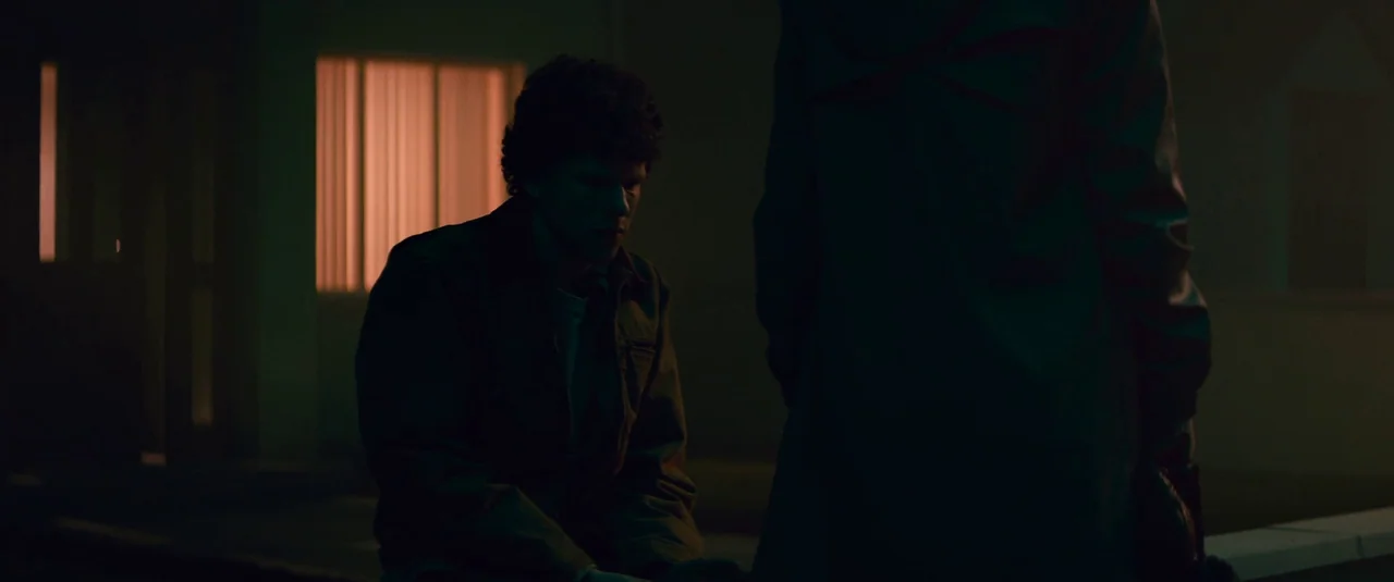 Vivarium (2019) movie still: medium — An exterior nighttime scene at a dimly lit building with a glowing orange window. A man sits on the curb while…