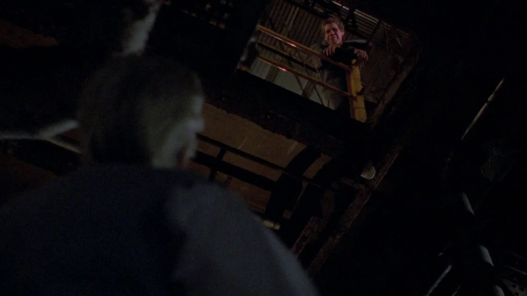 Cronos (1993) movie still: medium — An industrial metal walkway sits in near-darkness with yellow safety railing elements visible. A man stands on…