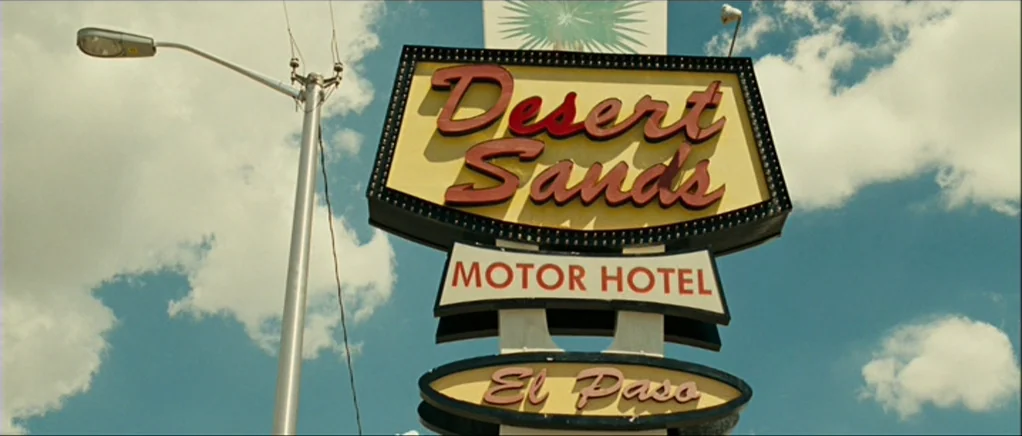 No Country For Old Men movie still: wide — A roadside motel sign for Desert Sands stands against a bright, cloudy blue sky. The metallic structure…