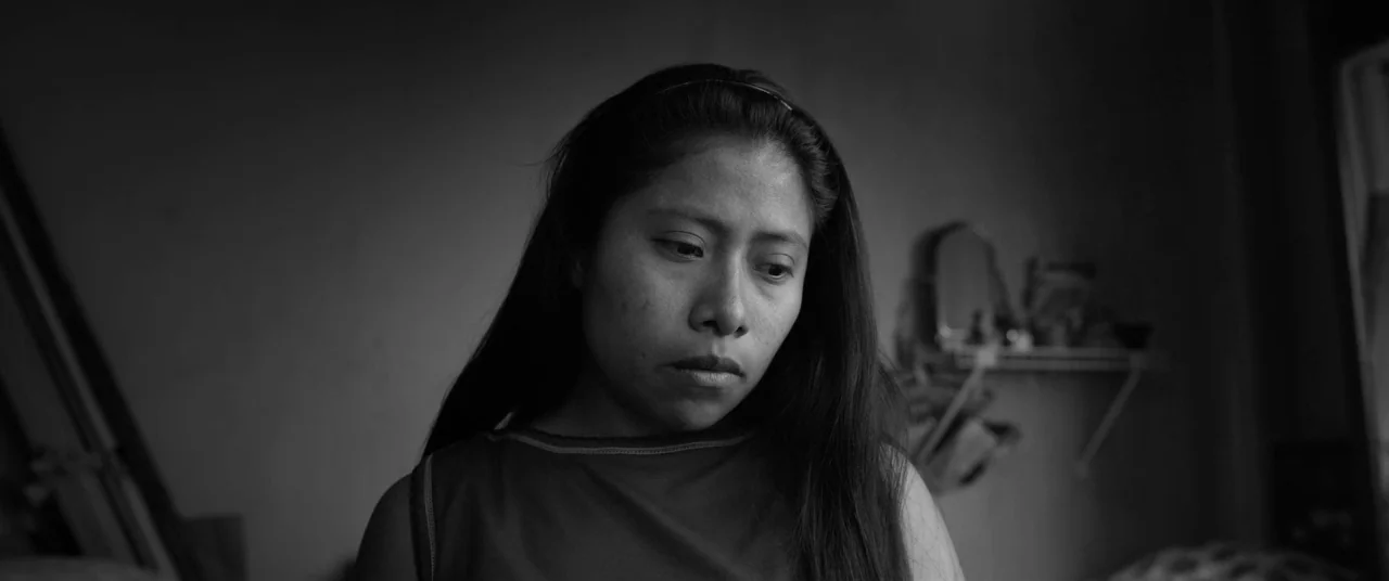 Roma (2018) movie still: closeup — A dimly lit room with a shelf and mirror blurred in the background. A young woman with long dark hair gazes…