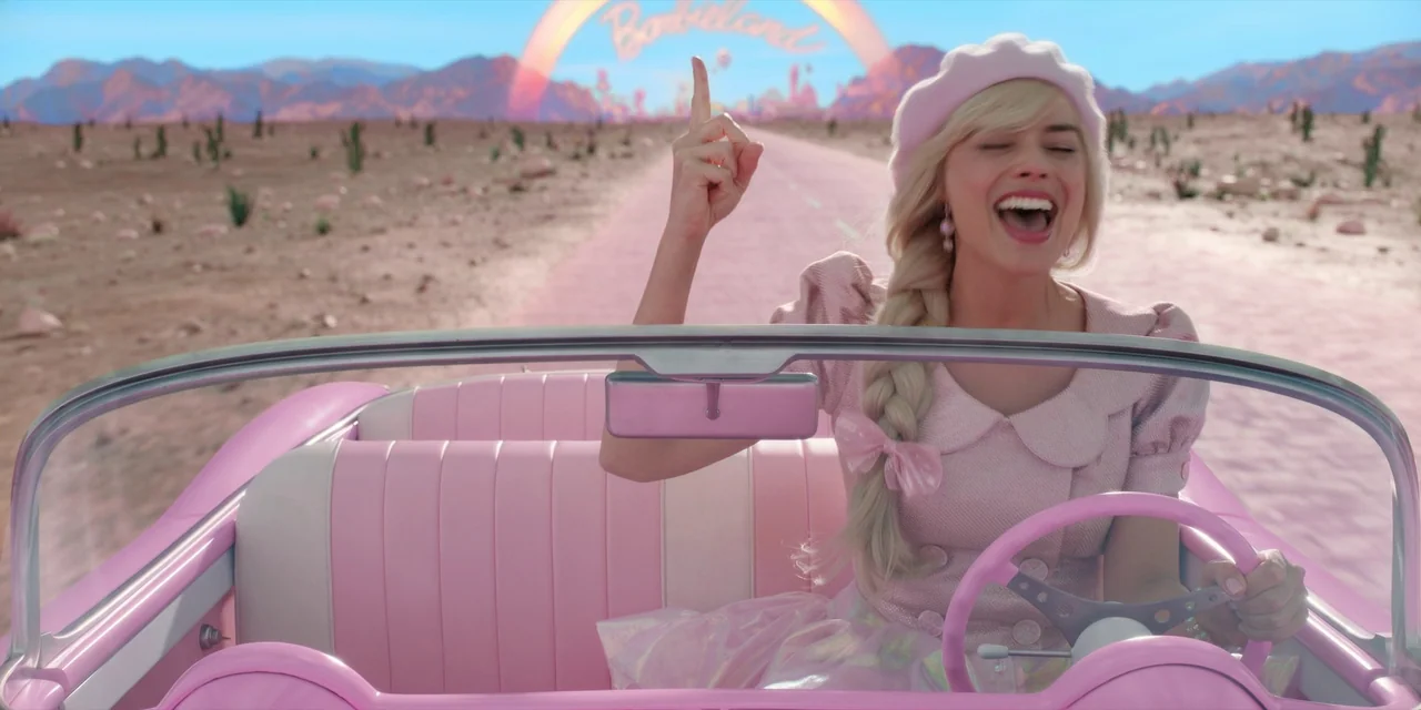 Barbie (2023) movie still: medium — A pink convertible drives down a desert road beneath a bright blue sky. A smiling blonde woman wearing a pink…