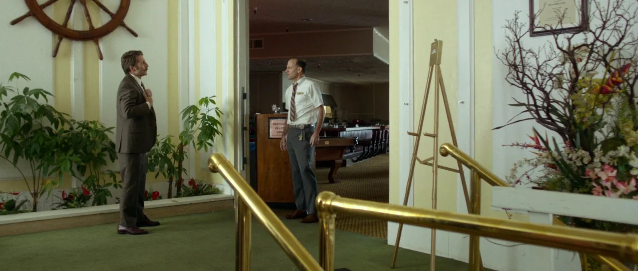 Faults (2014) movie still: wide — A vintage 1970s lobby with green carpet and a decorative wooden ship wheel. Two men in work attire stand facing…