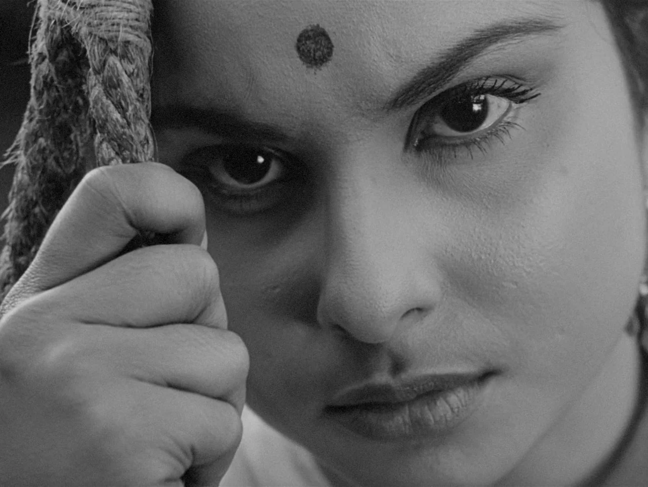 Charulata (1964) movie still: closeup — A close-up of a person's face set against a rustic background with rope texture. The person stares directly…